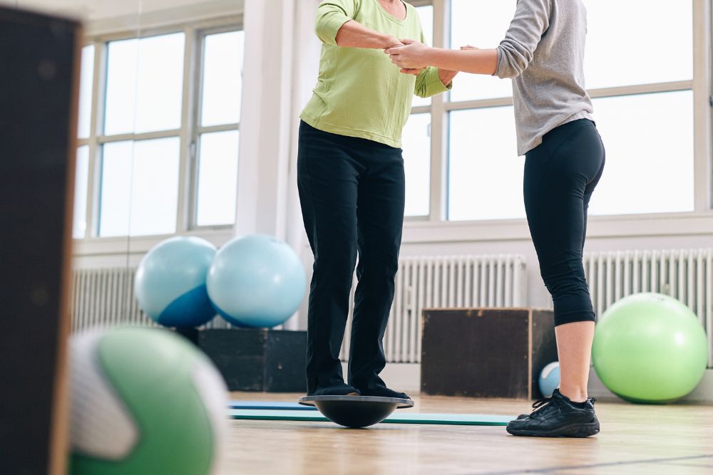 A physical therapist holding onto the hands of a woman balancing on a half sphere device as part of balance disorder treatment near Belfry, Blue Bell, Pennsylvania.